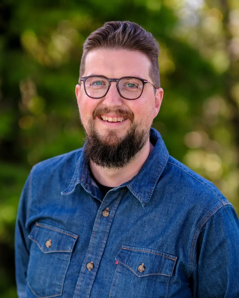 En mann med briller og skjegg smilende foran trær.